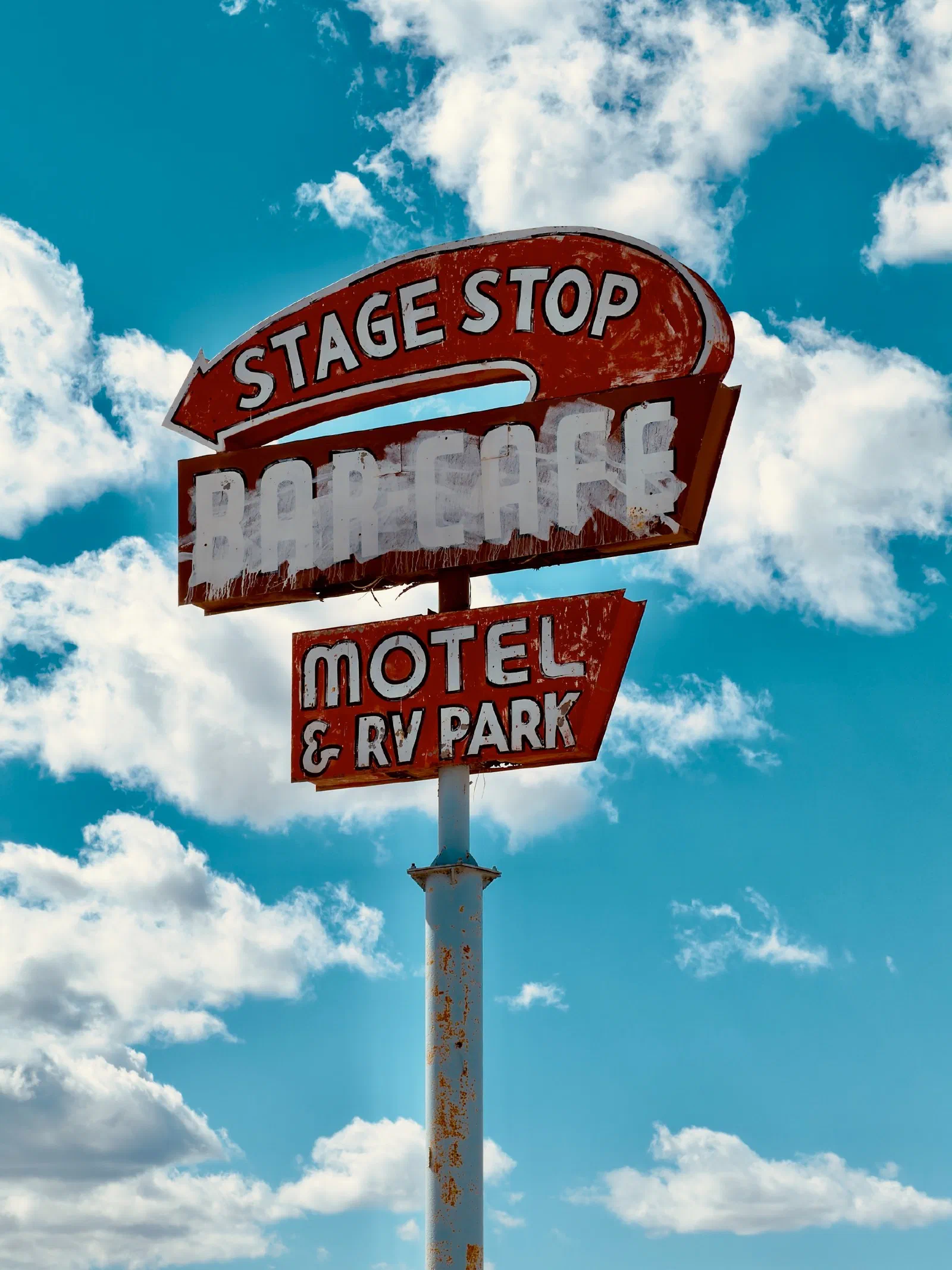 A vintage red sign for a motel and RV park with the words Stage Stop against a blue sky with scattered clouds.
