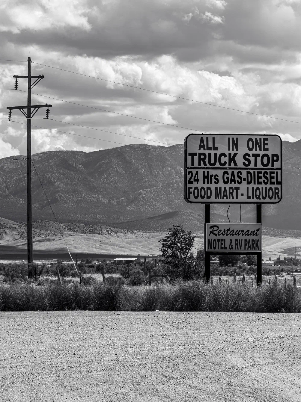 A roadside sign advertises an all-in-one truck stop offering 24-hour gas, diesel, food mart, liquor, restaurant, motel, and RV park services against a mountainous backdrop.
