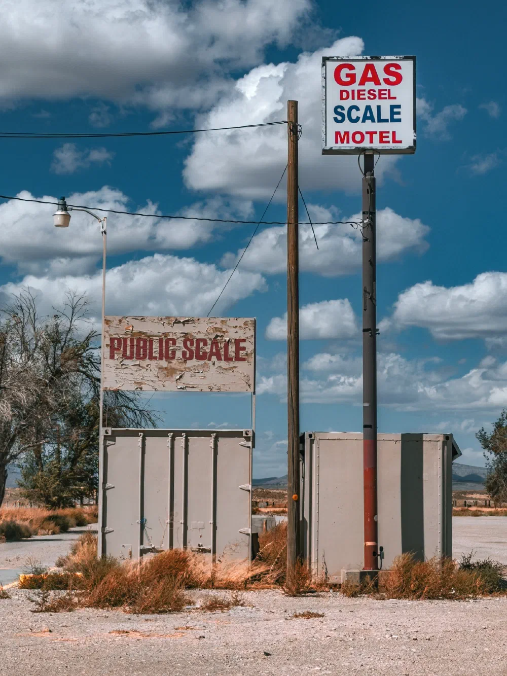 A roadside setup displays signs for gas, diesel, scale, motel, and a weathered public scale in a rural area under a partly cloudy sky.