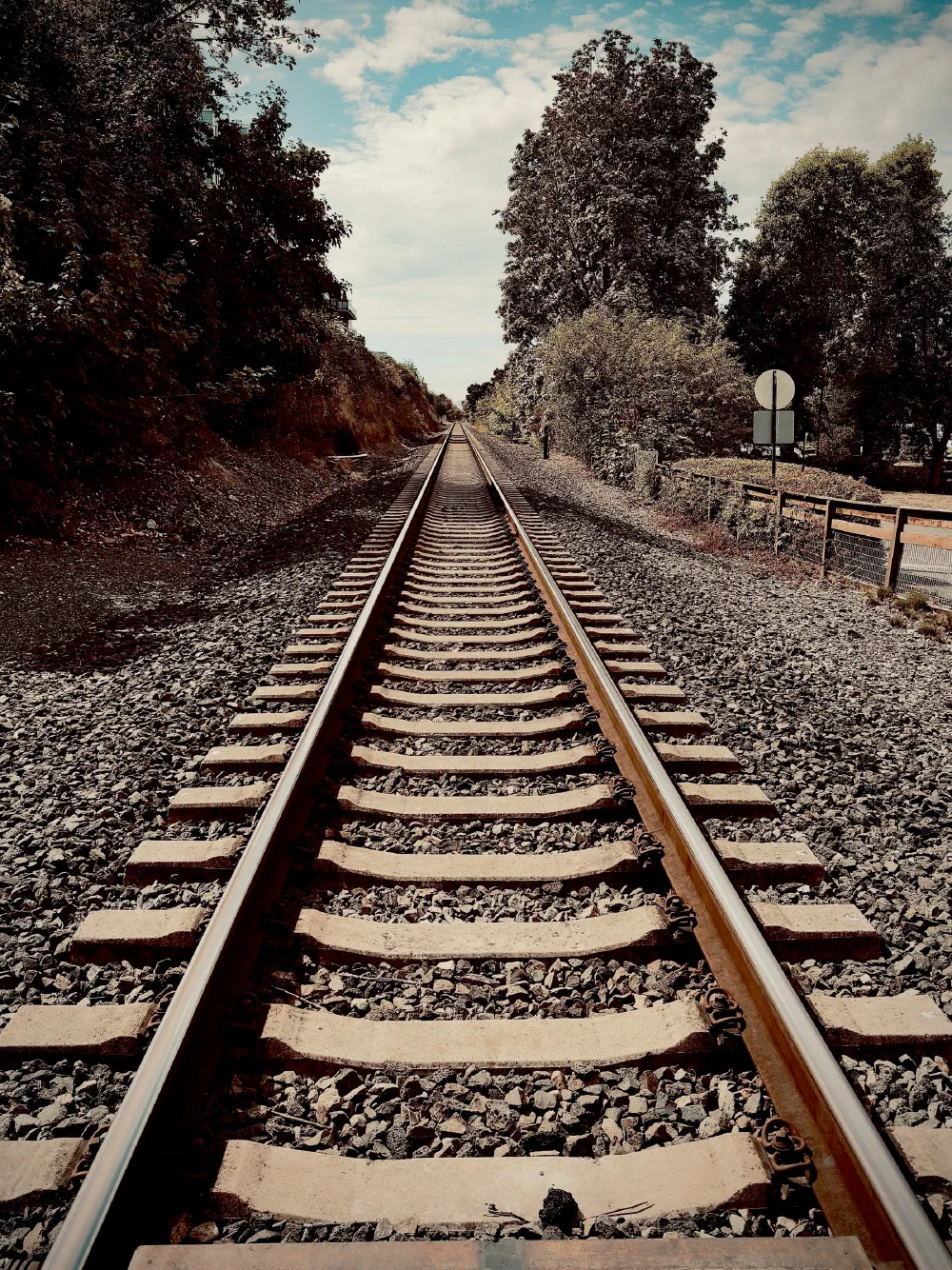 A single set of railroad tracks extends straight into the distance, flanked by trees and gravel on either side under a partly cloudy sky.