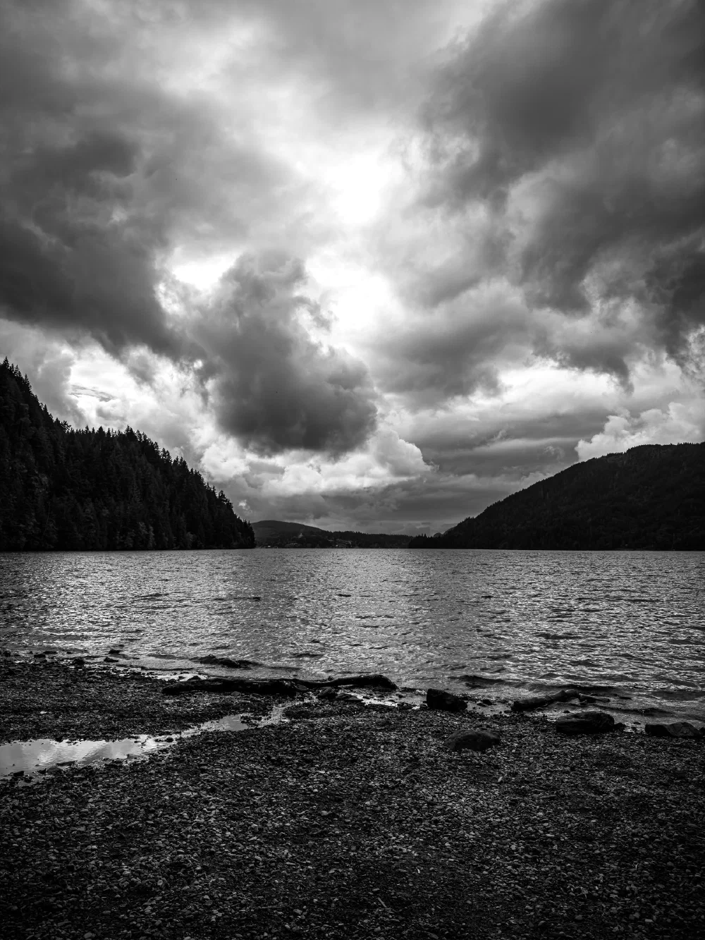 A serene lake is surrounded by tree-covered hills under a dramatic, cloudy sky.
