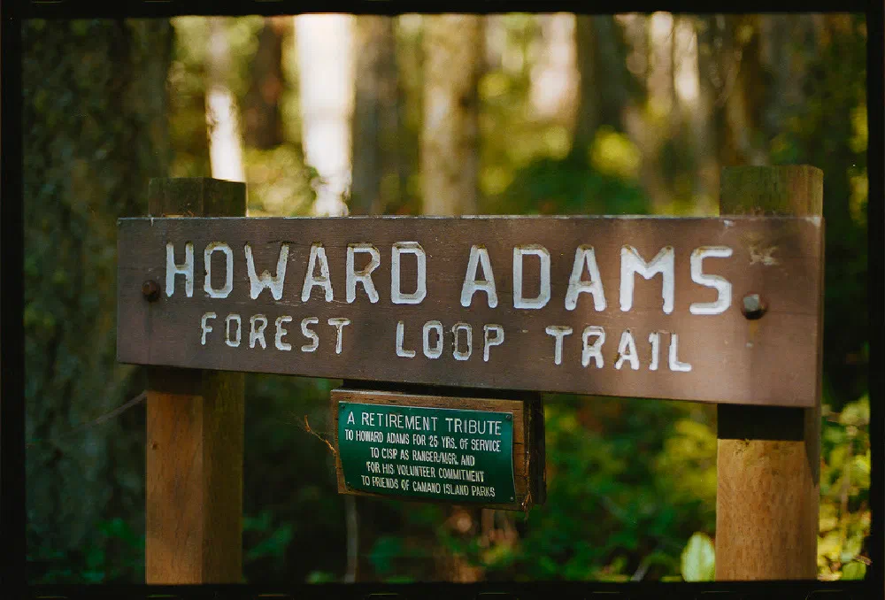 A wooden sign marking the Howard Adams Forest Loop Trail stands in a wooded area, accompanied by a smaller plaque honoring Howard Adams for his service and volunteer commitment.