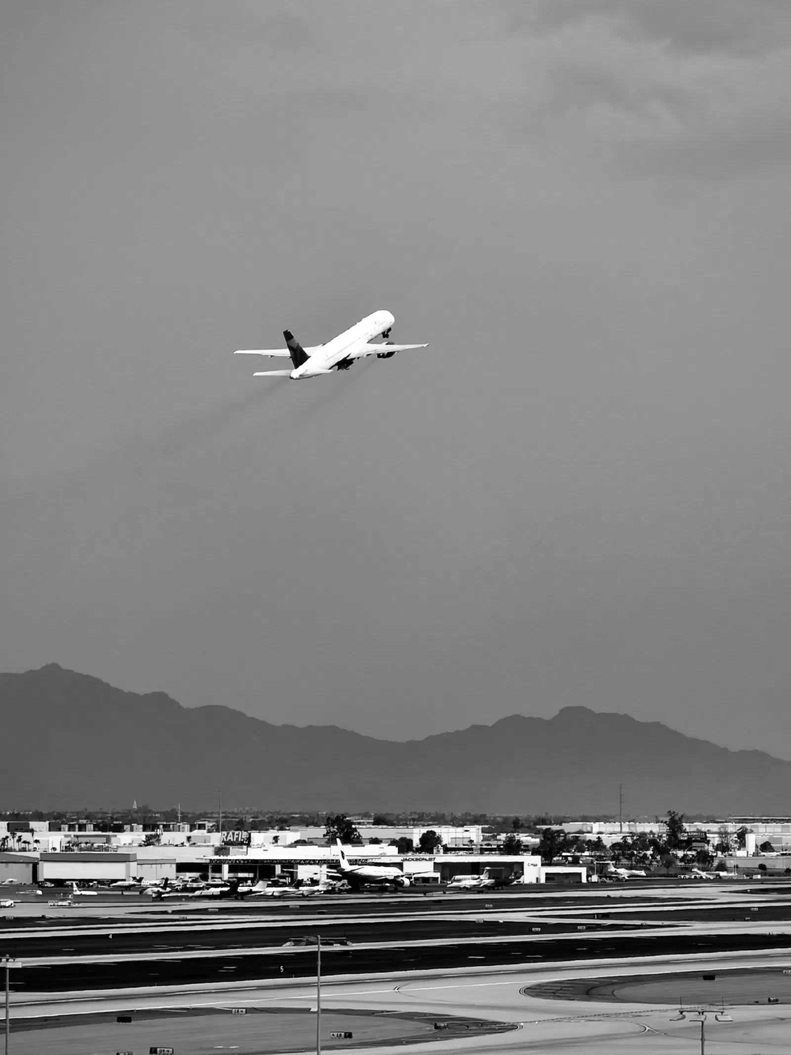 An airplane is ascending into the sky above an airport runway with mountains in the background.