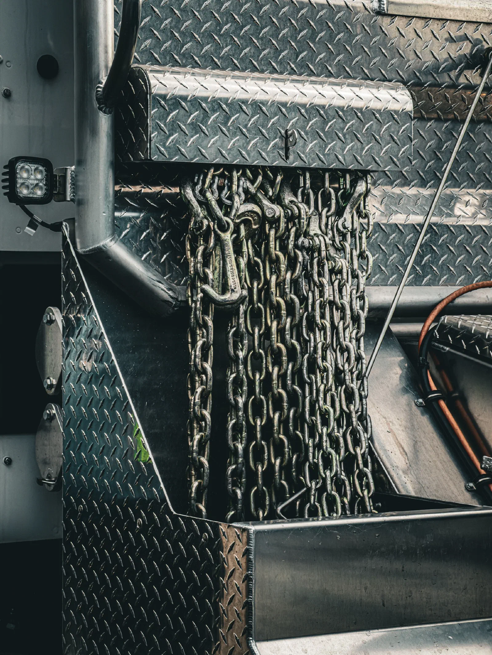 Heavy metal chains with hooks are hanging from a compartment on a diamond plate metal surface.