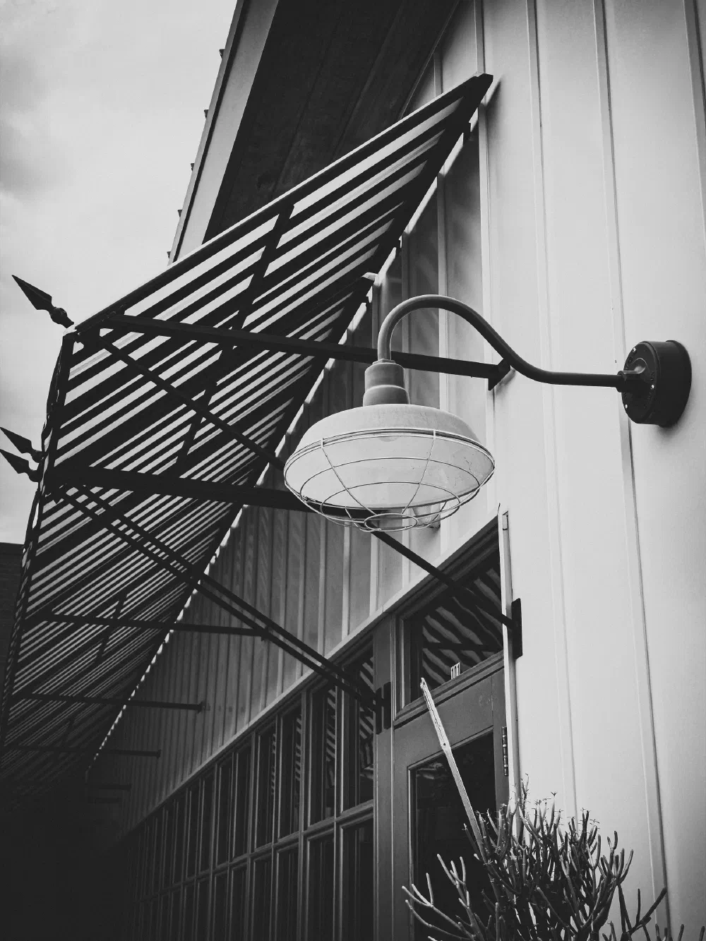 A black and white photo showcases an exterior wall with a vintage industrial-style light fixture and a metal awning above a door and windows.