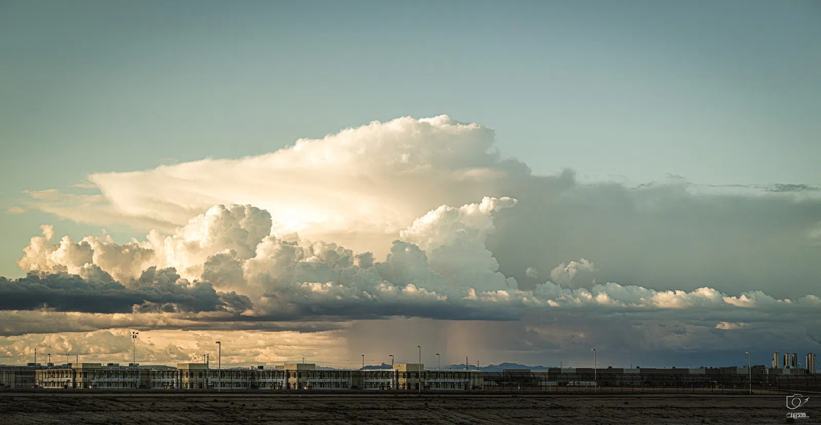 A dramatic sky filled with large, dense clouds hovering over a distant industrial or warehouse complex near a barren landscape.